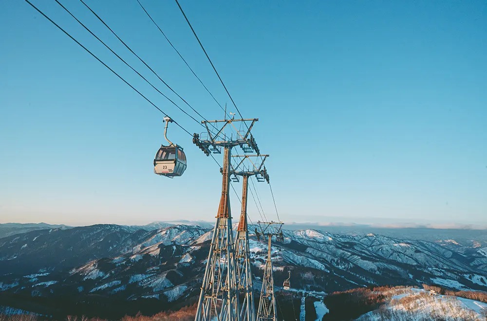 青空の下、雪をかぶった山並みの上を進むゴンドラと鉄塔