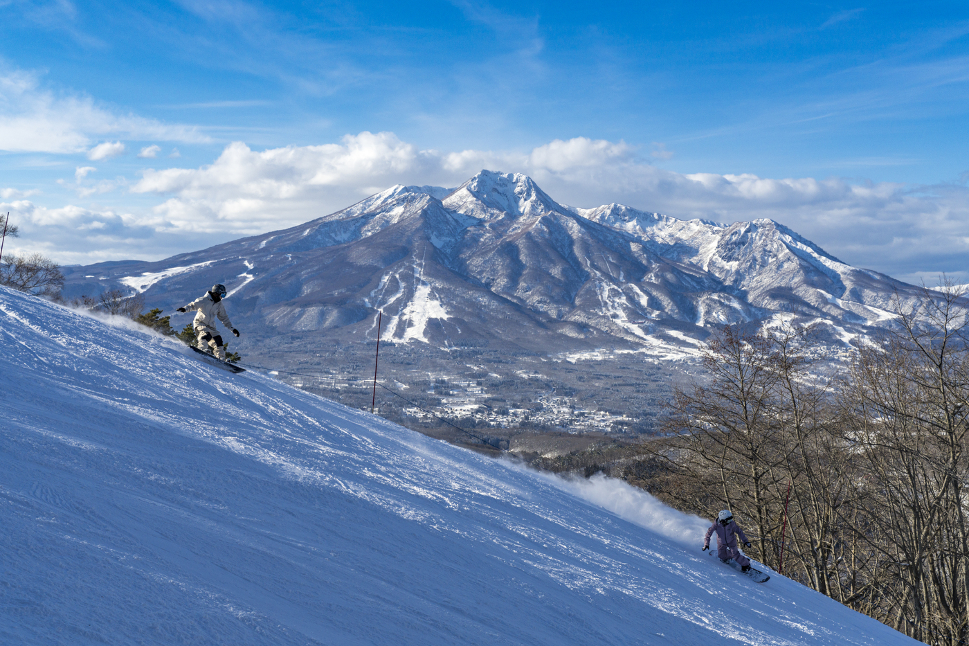 雪の山々を正面に見ながら急斜面を滑り降りるスノーボーダー2人
